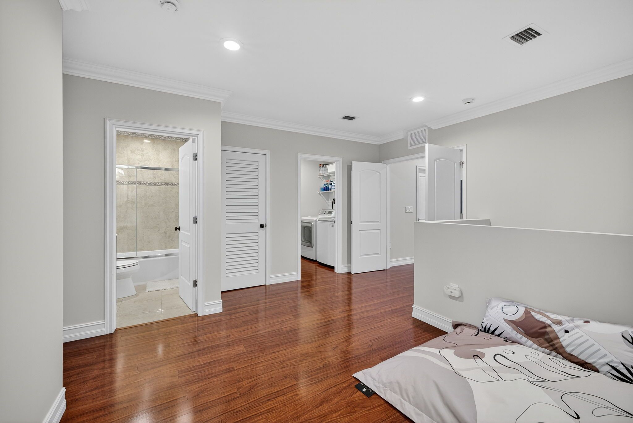A bedroom with a wooden floor, a bed with patterned bedding, a white wardrobe, and a bathroom with a shower and toilet.