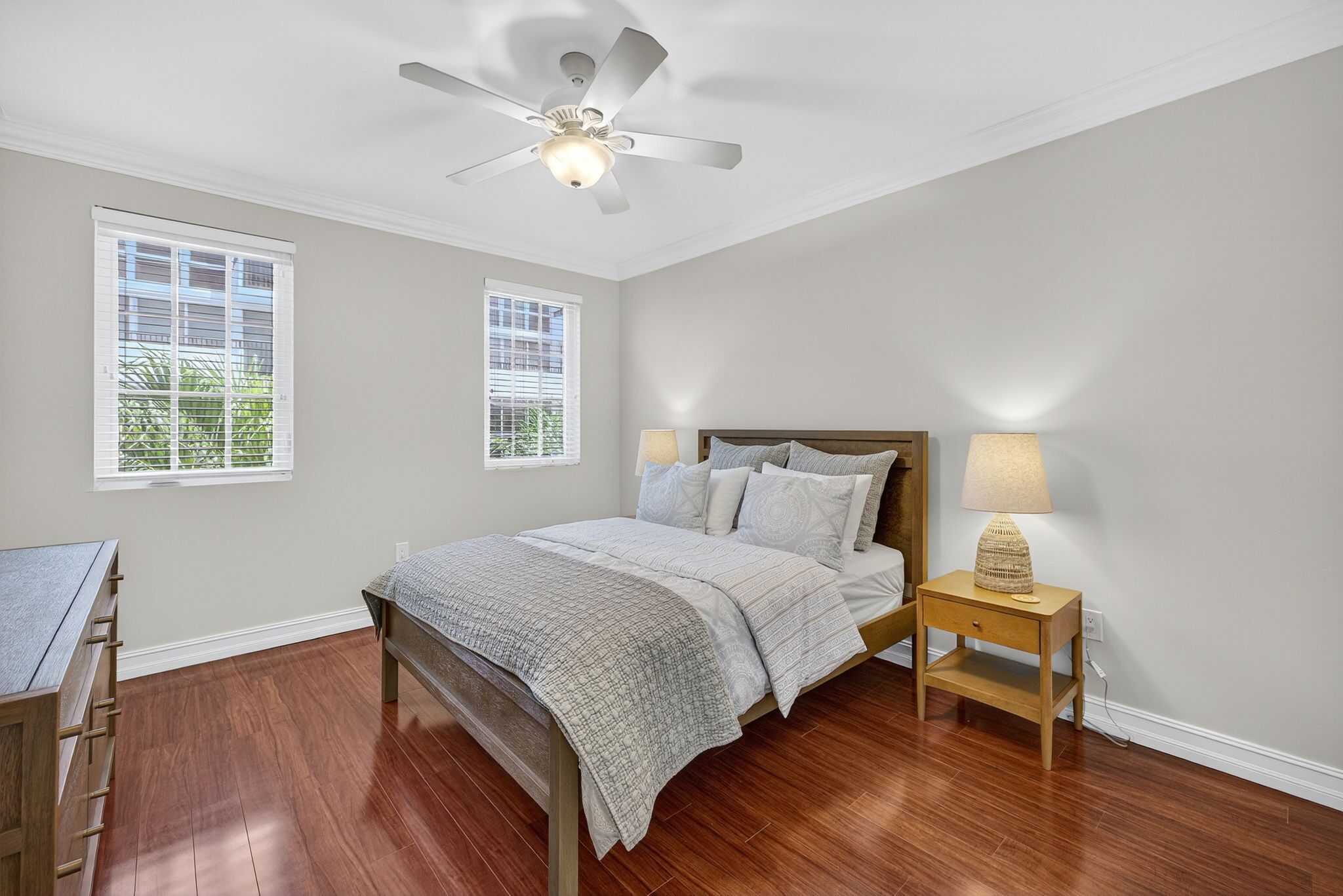 A bedroom with a bed, two bedside tables with lamps, a ceiling fan, and two windows.