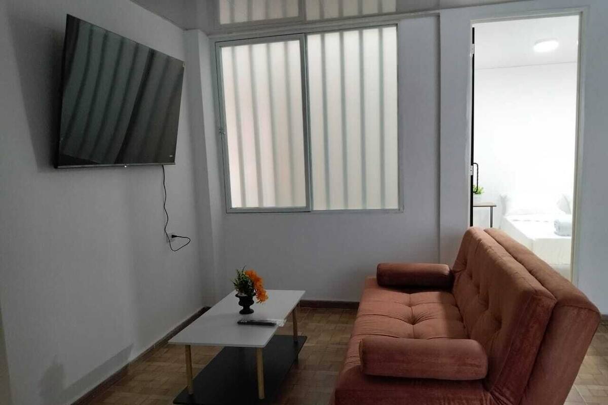 A living room with a flat-screen TV mounted on the wall, a brown sofa, a small table with a vase of flowers, and large windows with blinds.