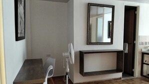 Una stanza con pavimento in legno, una sedia bianca, un tavolino, una ventola da terra, uno specchio e un mobile consolle scuro.