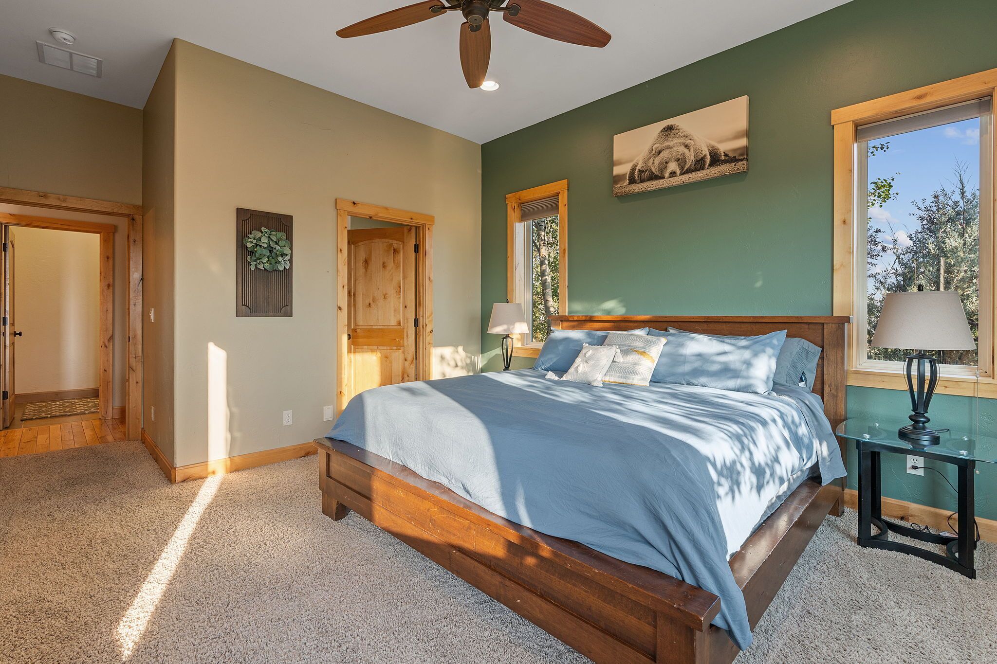 Une chambre avec un grand lit, une tête de lit en bois, un ventilateur de plafond, une fenêtre avec vue et une porte donnant sur une autre pièce.