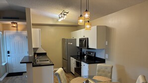A compact kitchen with a refrigerator, microwave, and stove. There is a dining table with chairs and a ceiling light fixture. - Peaceful 2-bedroom condo at the beach with a water view! (Surfside Beach)
