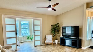 A living room with a beige sofa, a TV on a black stand, a ceiling fan, and a large window with a view of a house. - Peaceful 2-bedroom condo at the beach with a water view! (Surfside Beach)