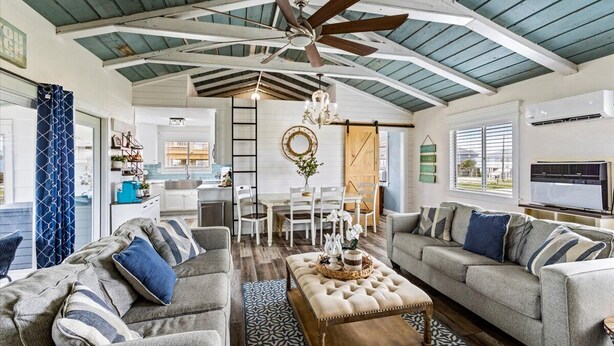 A spacious living room with a high ceiling, a ceiling fan, and a large window. There is a dining area with a table and chairs, and a kitchen in the background. - SAN LUIS BEACH GEM - Bay & Gulf Views, Boat launch access, Short Walk to Beach (Freeport)