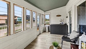 A covered porch with a wooden floor, a black wicker chair, a small potted plant, a trash can, and a dark-colored cabinet. - SAN LUIS BEACH GEM - Bay & Gulf Views, Boat launch access, Short Walk to Beach (Freeport)
