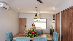 A modern dining area with a wooden table and blue upholstered chairs, a kitchen with a refrigerator and stove, and a large window. - Magical oasis perfect: Family, surf, relax and nature, close to Tamarindo beach (Tamarindo)