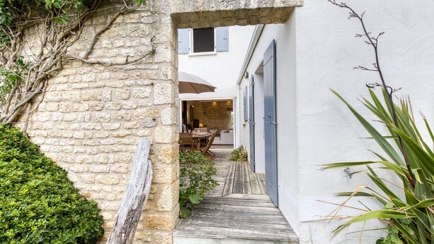 A narrow stone pathway leading to a wooden deck with a table and chairs, surrounded by greenery and a white building with blue shutters. - Gorgeous home in Saint-Pierre-d'Oléron with WiFi (Saint-Pierre-d'Oléron)