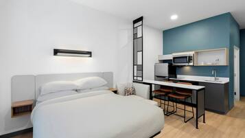 A modern hotel room with a bed, bedside table, and a kitchenette area including a microwave and bar stools.