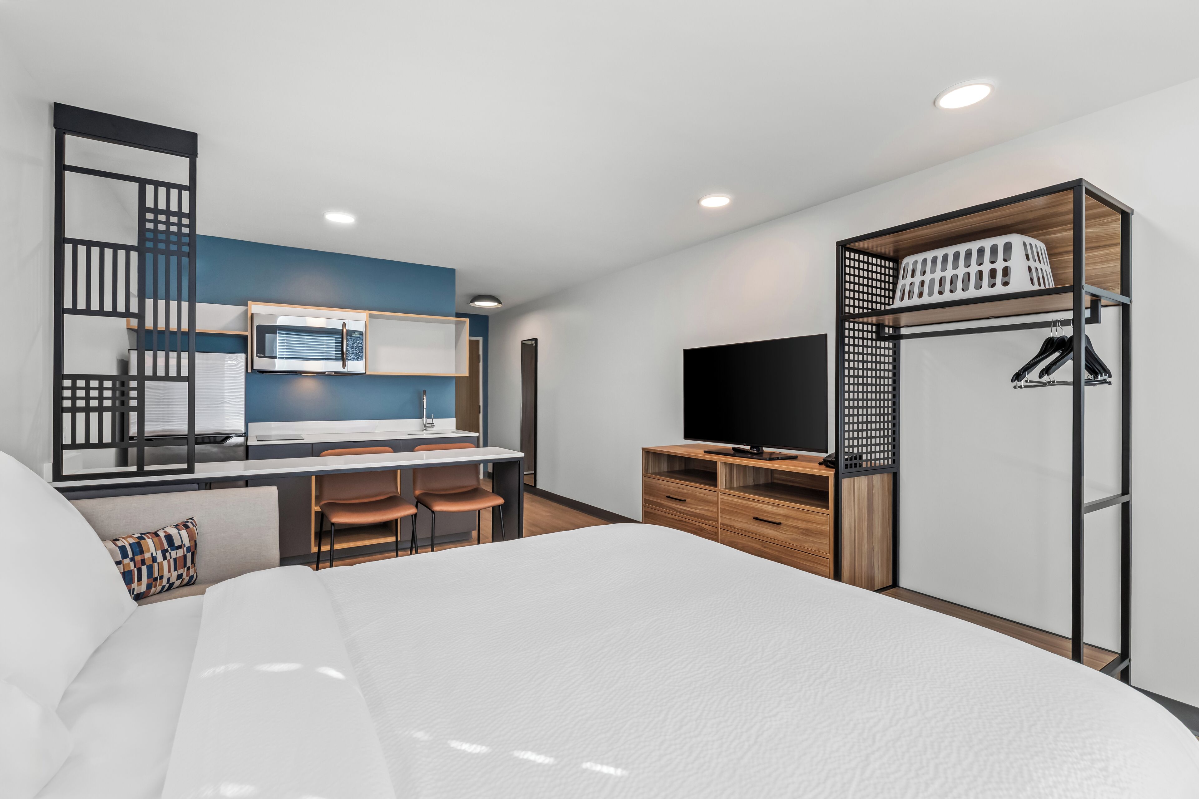 A modern hotel room with a bed, a desk with a chair, a TV, a wardrobe, and a kitchenette.