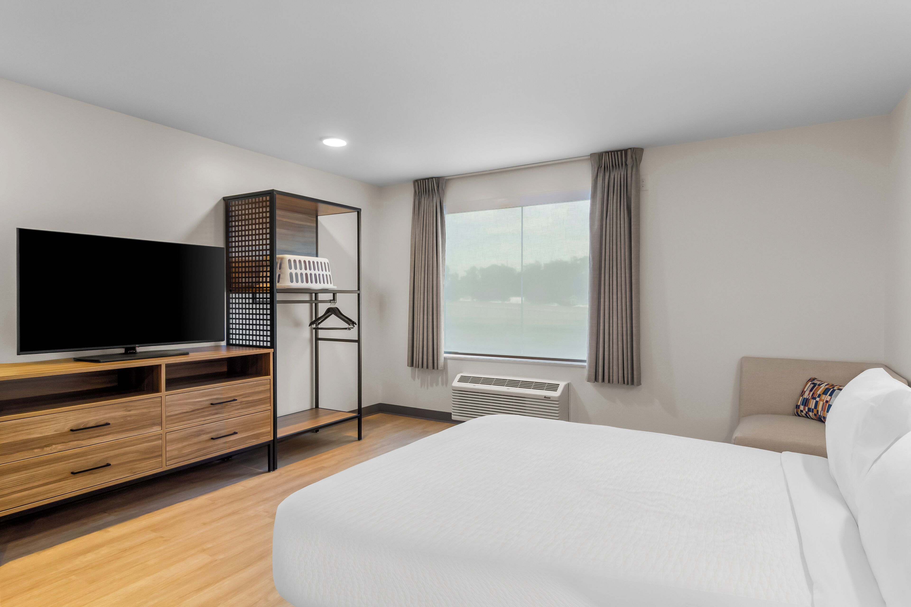 A hotel room with a bed, a TV, a wooden dresser, a hanging clothes rack, and a window with curtains.