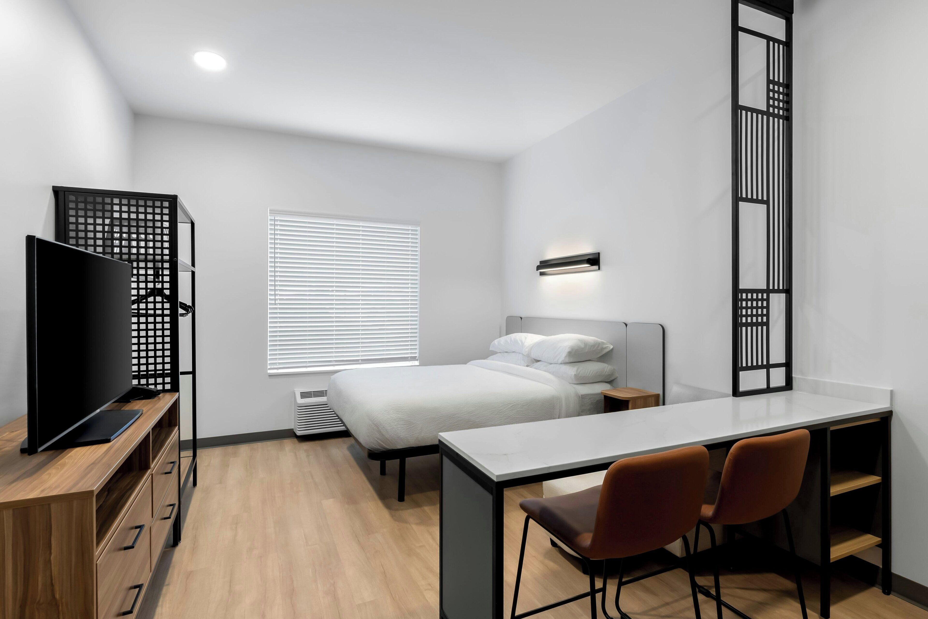 A modern, compact room with a bed, a desk, two chairs, a TV, and a window with blinds.