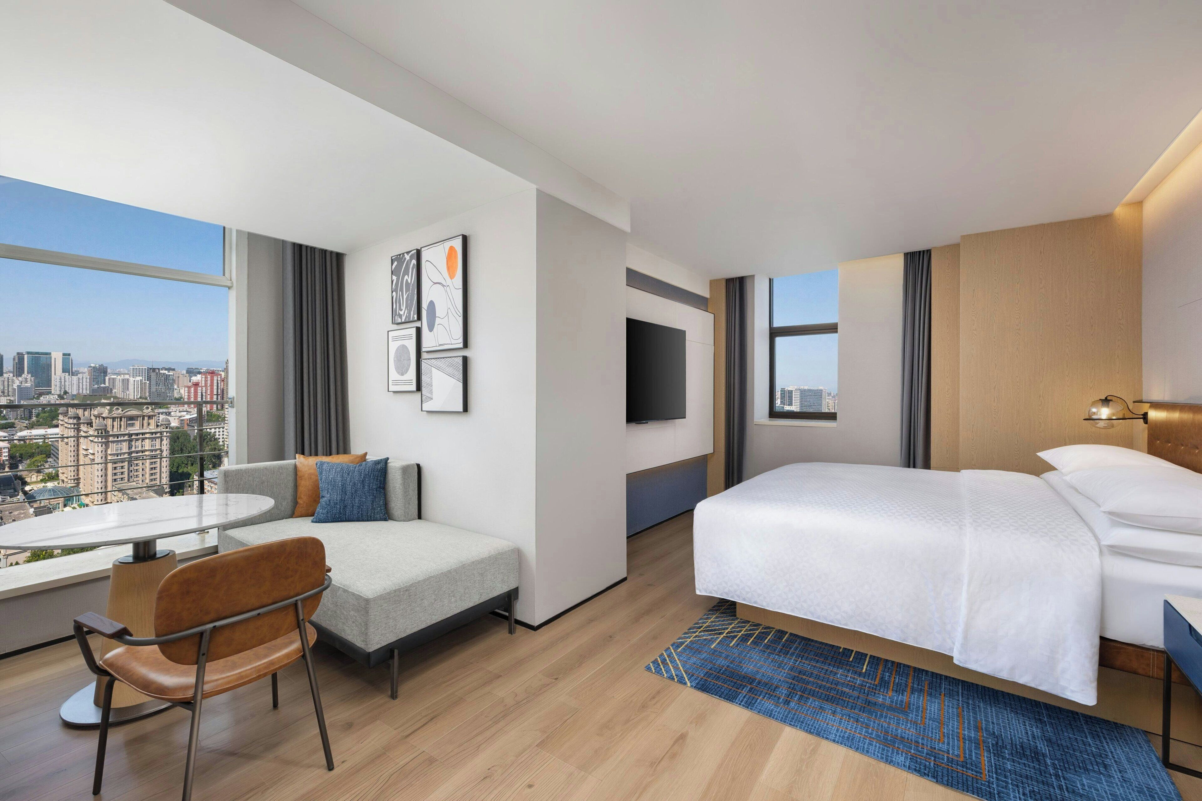 A modern hotel room with a large bed, a sofa, a dining table, and a TV. The room has a city view.
