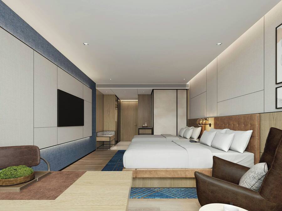 A modern hotel room with a large bed, a seating area with a brown armchair, a small table with a plant, and a TV mounted on the wall.