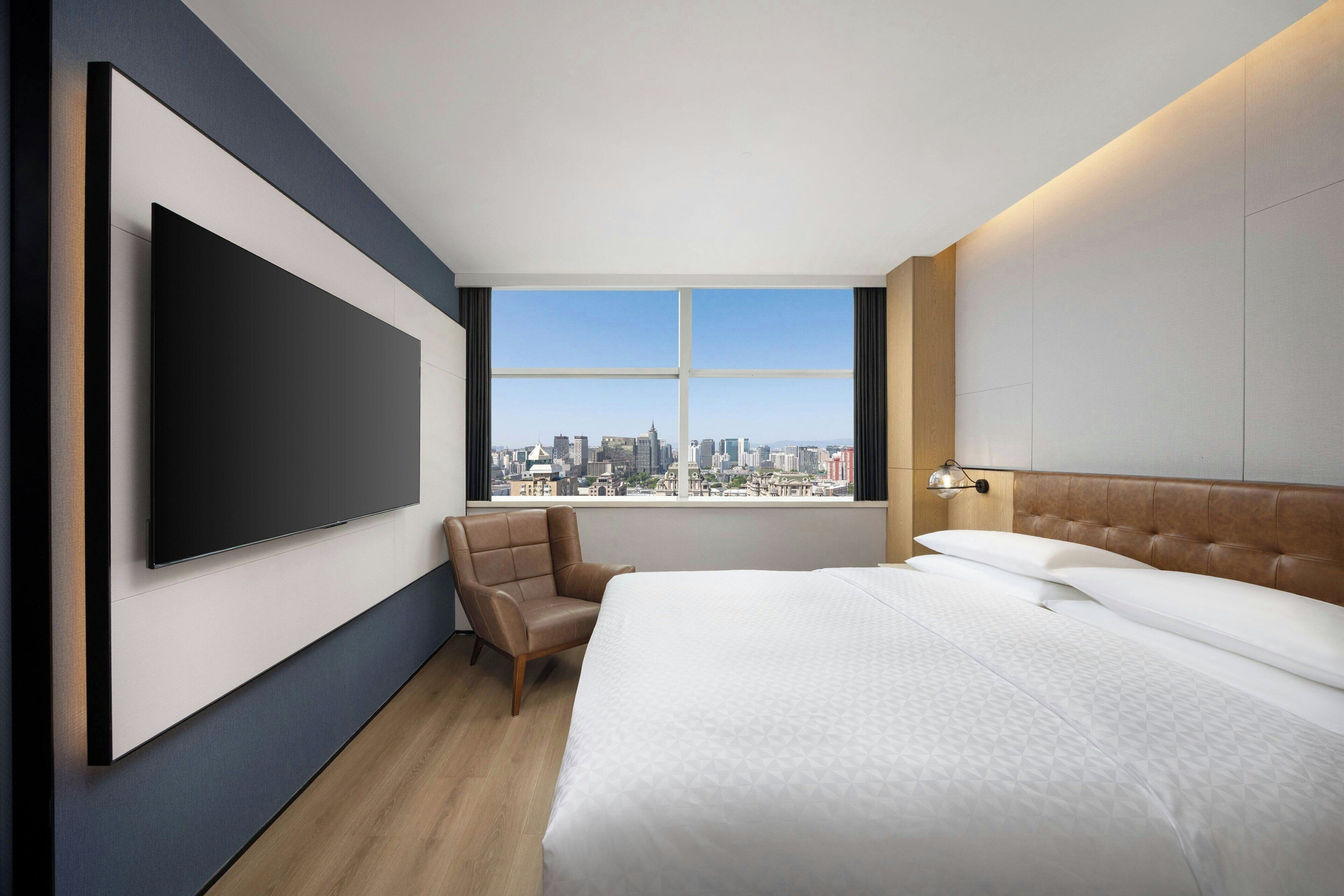 Una habitación de hotel moderna con una cama grande, una silla, un televisor de pantalla plana en la pared y vistas a la ciudad desde la ventana.