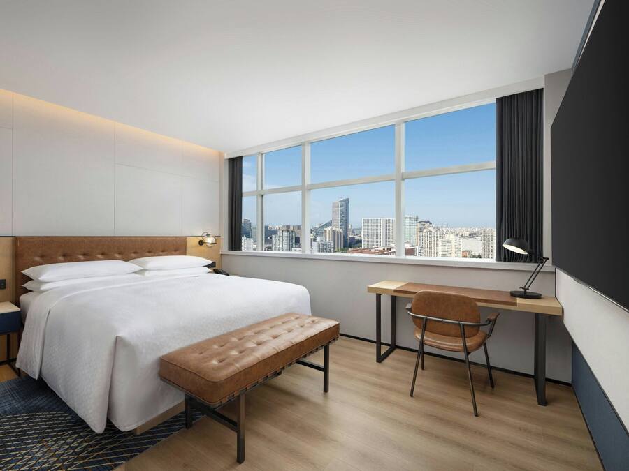 A modern hotel room with a large bed, a desk, and a view of the city skyline.