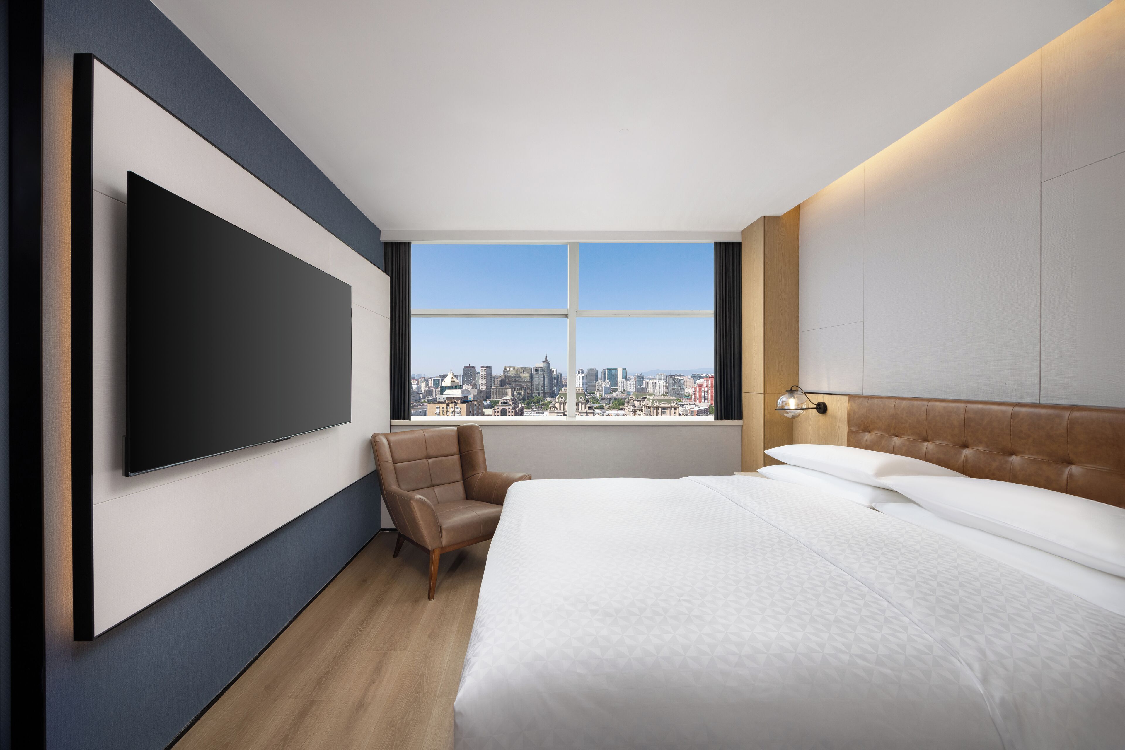 A modern hotel room with a large bed, a flat-screen TV mounted on the wall, a city view, and a leather armchair.