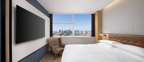 Una habitación de hotel moderna con una cama grande, un televisor de pantalla plana en la pared, vistas a la ciudad y un sillón de cuero.