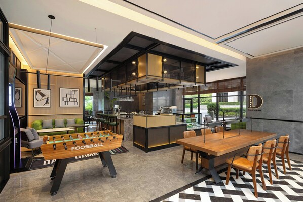 A modern hotel lobby with a foosball table, a bar area, and a dining space with a large wooden table and chairs.