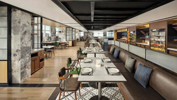 A modern restaurant with a long communal dining table, booths, and a counter with a display of food items. - Four Points By Sheraton Beijing Sanlitun Workers Stadium (Beijing)