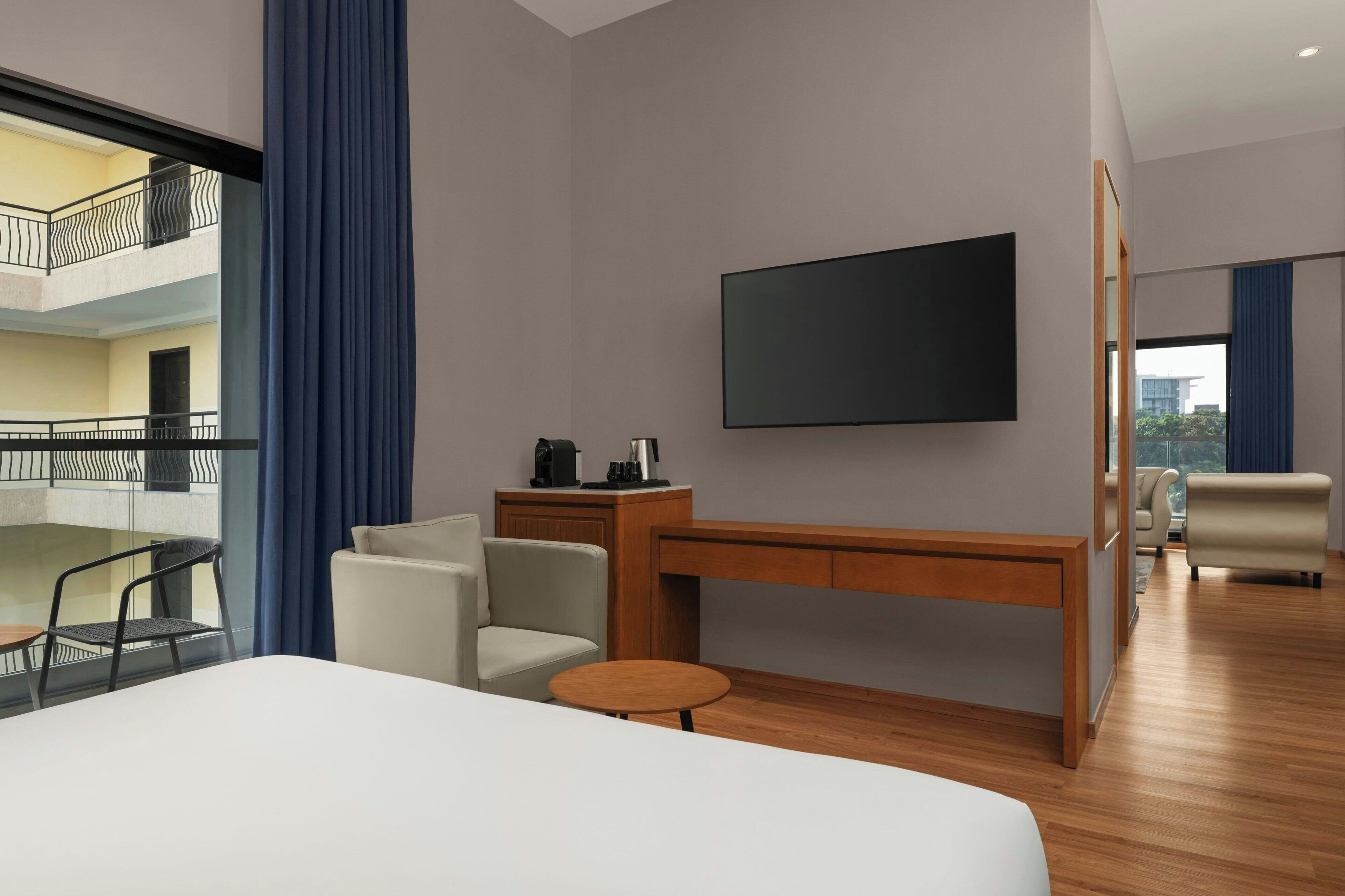 A hotel room with a bed, a desk, a chair, a small table, a TV, and a balcony with a railing.