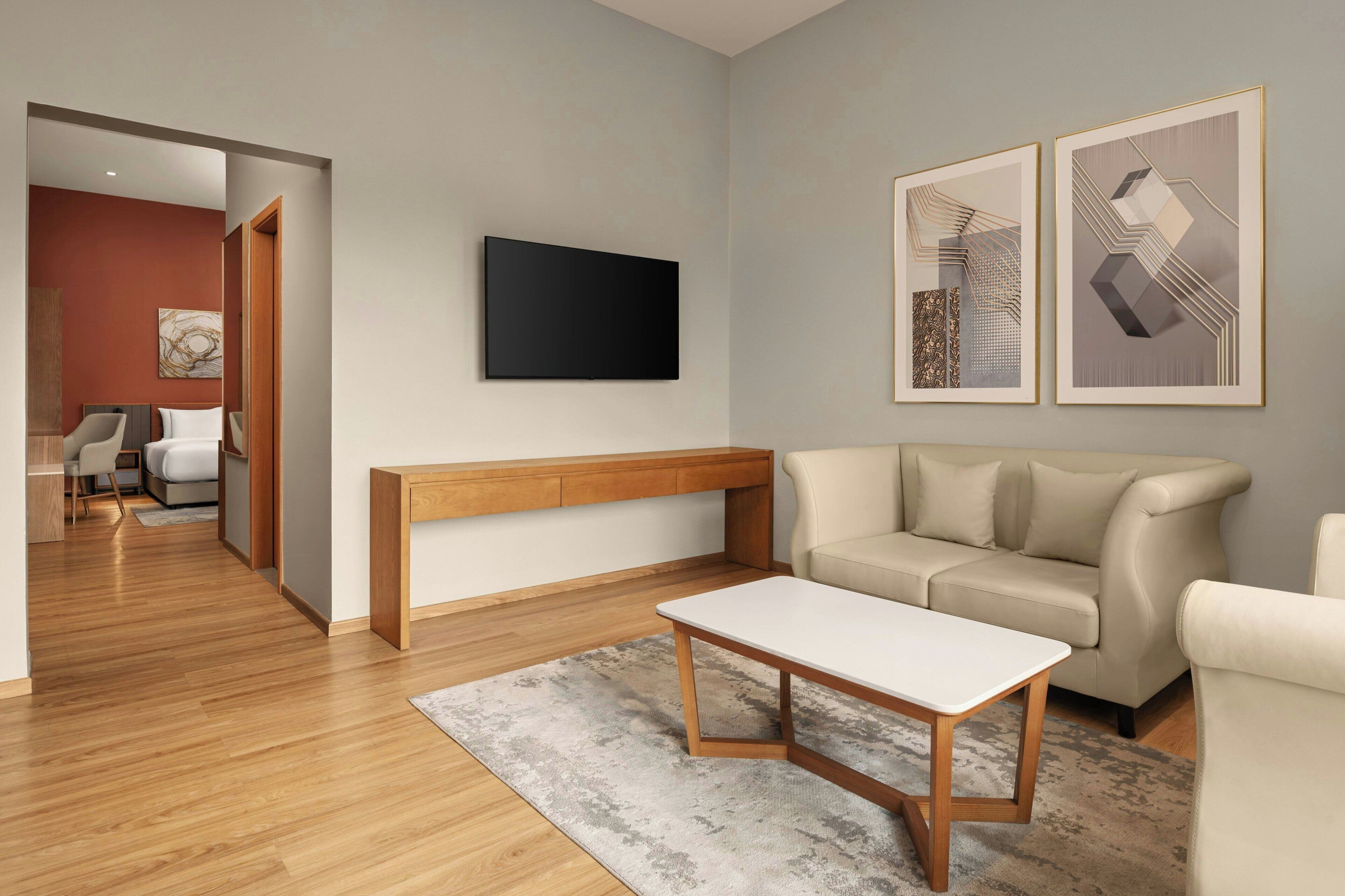 A modern living room with a wooden bench, a white coffee table, a beige sofa, and a flat-screen TV mounted on the wall.