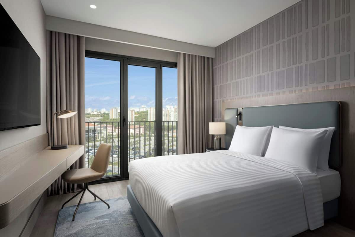 Habitación de hotel con cama, escritorio, silla y un amplio ventanal con vista a la ciudad.