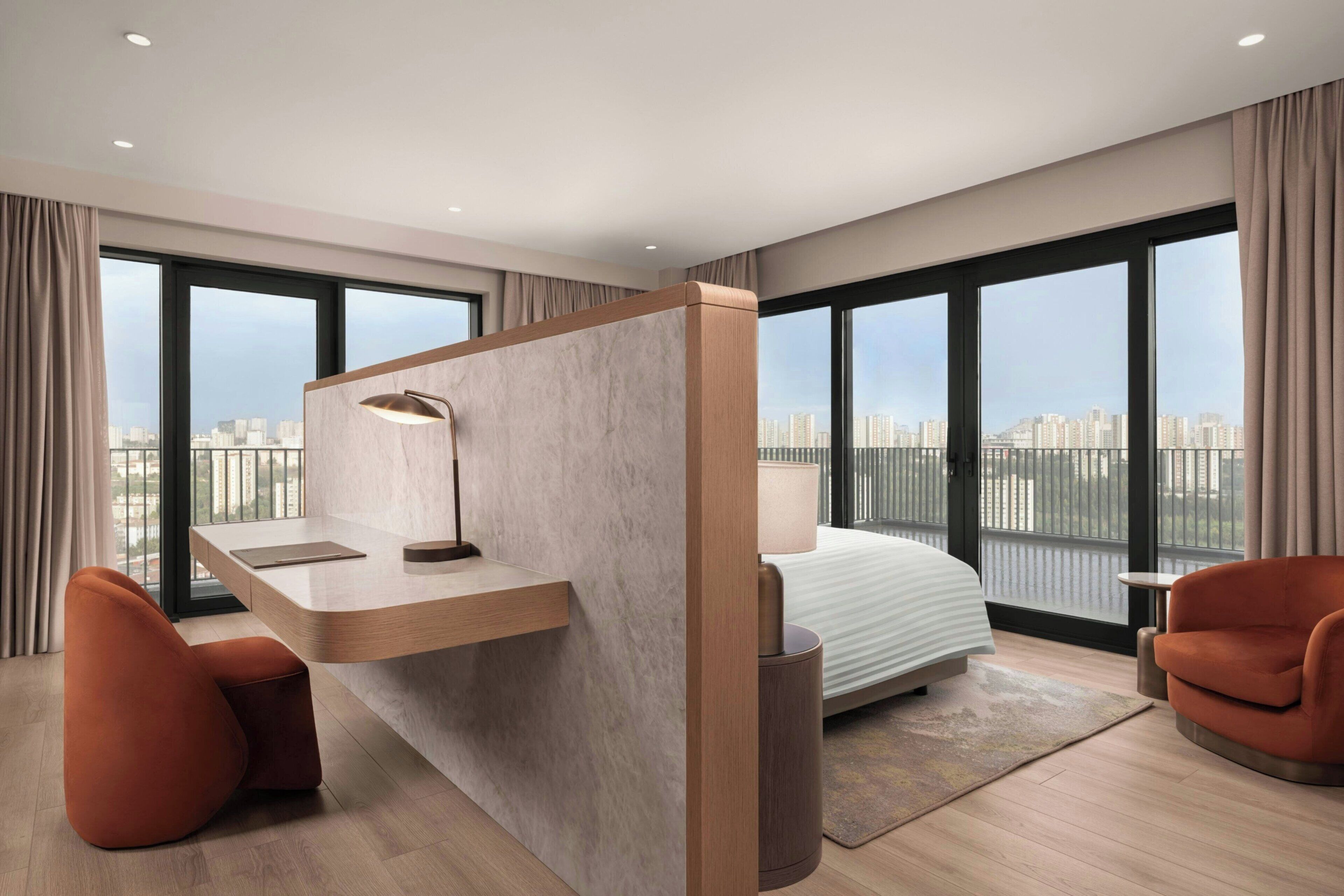 Quarto de hotel moderno com uma cama grande, uma escrivaninha com abajur e vista para a cidade.