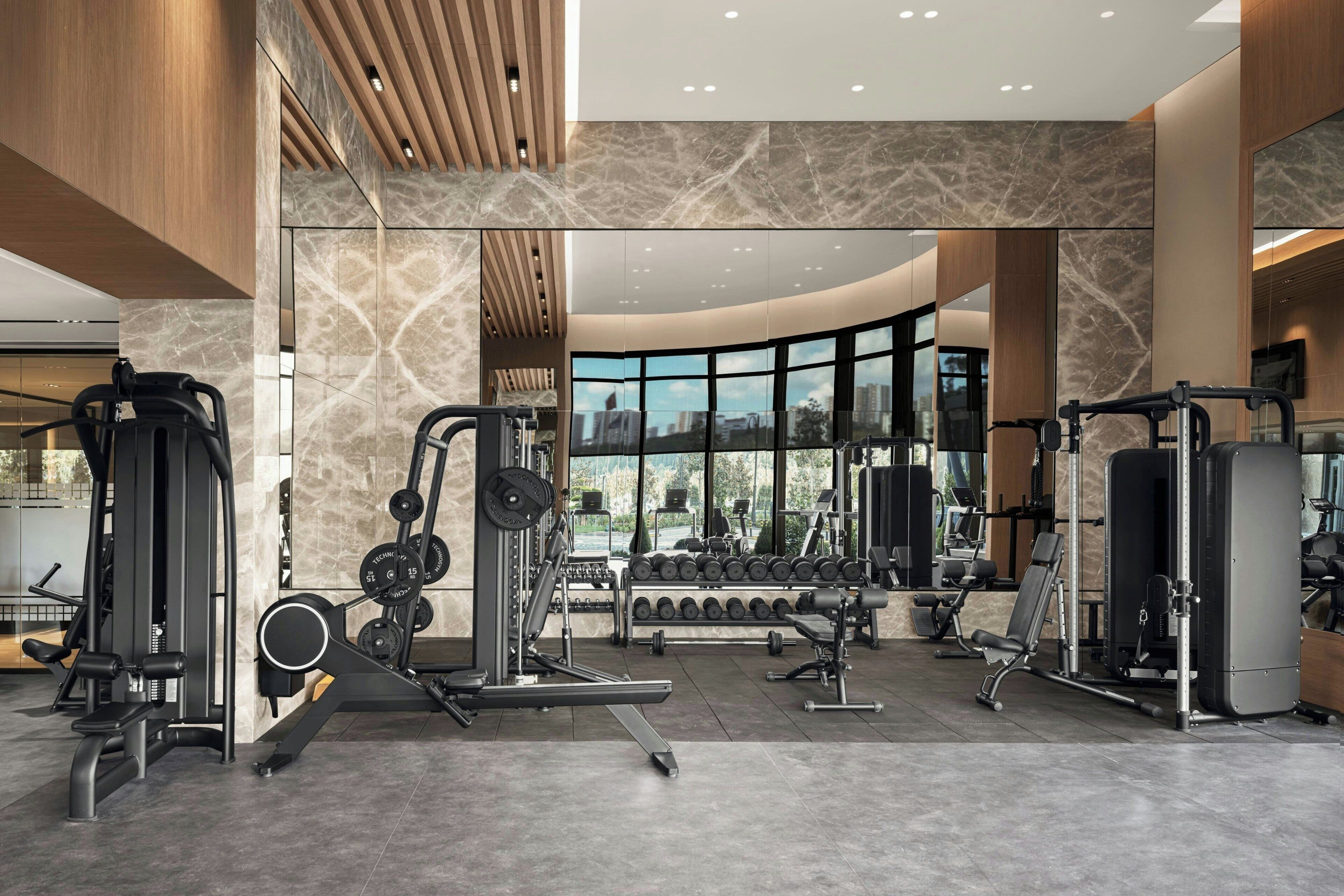A modern gym with various exercise equipment, large windows, and a marble floor.