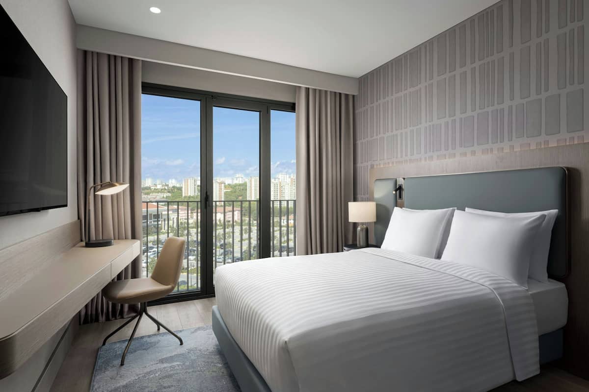 Habitación de hotel con una cama grande, un escritorio con una silla, un televisor y vistas a la ciudad desde un balcón.