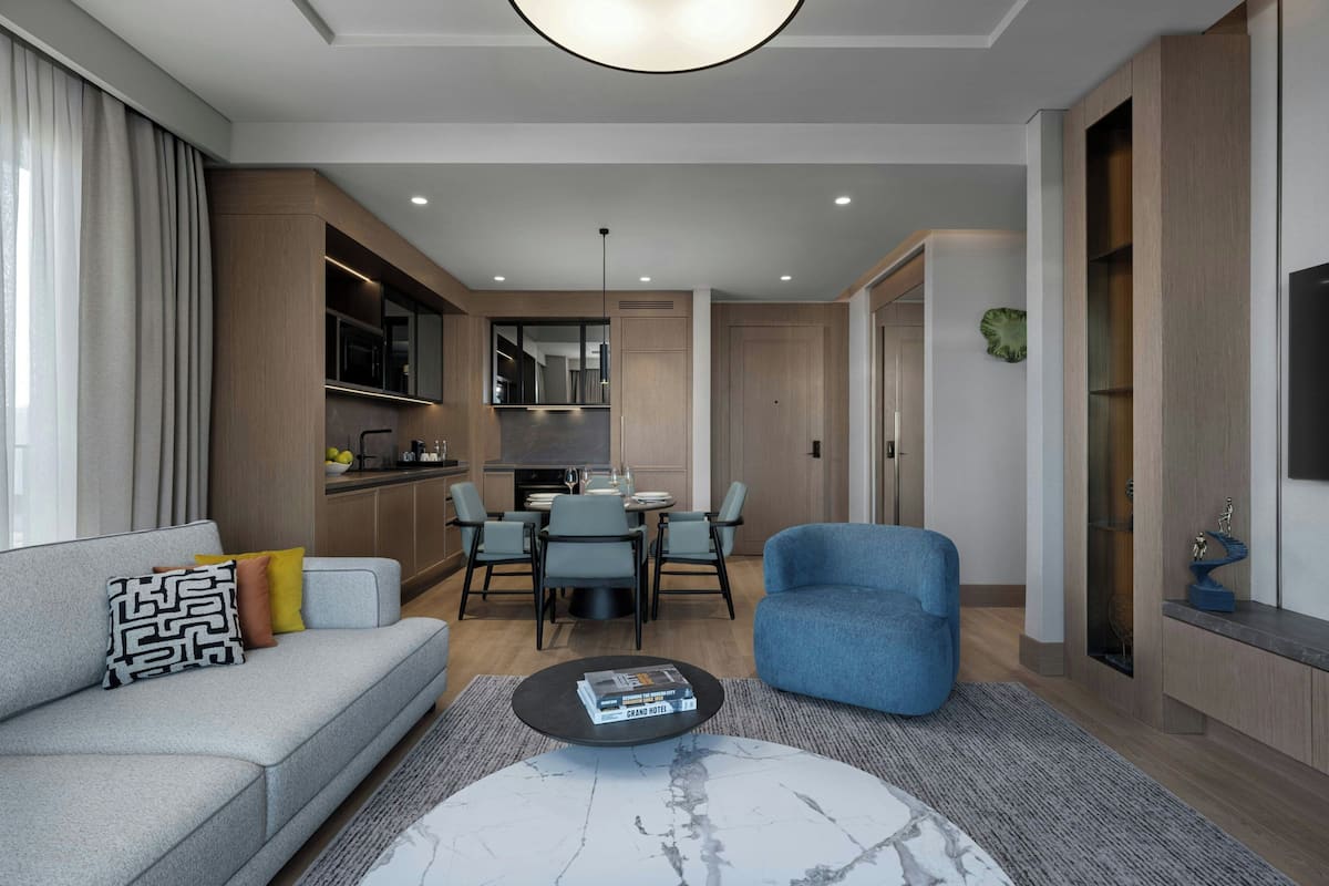 Una sala de estar moderna con un sofá gris, un sillón azul y una mesa de centro con libros. Se observa un comedor con mesa y sillas, y una cocina al fondo.