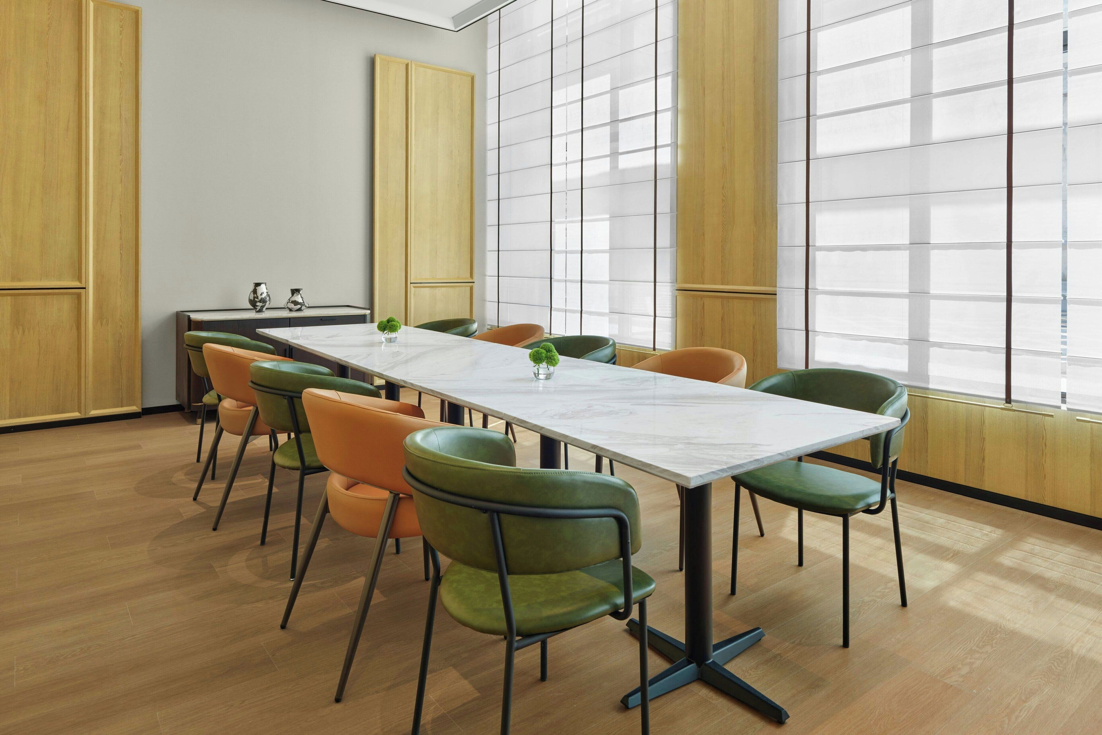 Un salón de conferencias con una larga mesa, sillas verdes y naranja, y ventanas amplias con persianas.