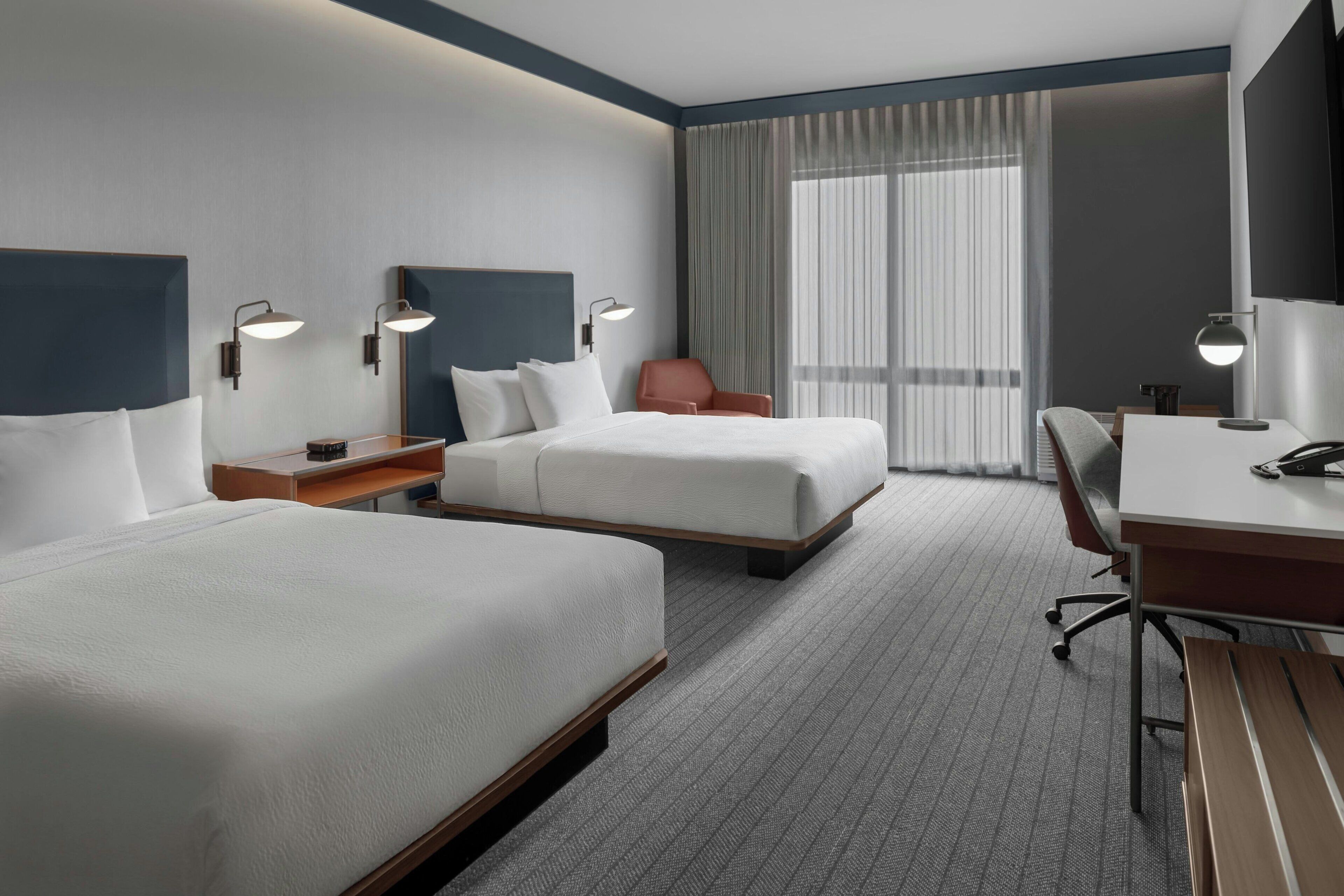 Una habitación de hotel con dos camas, un escritorio, una silla y un ventanal con cortinas.