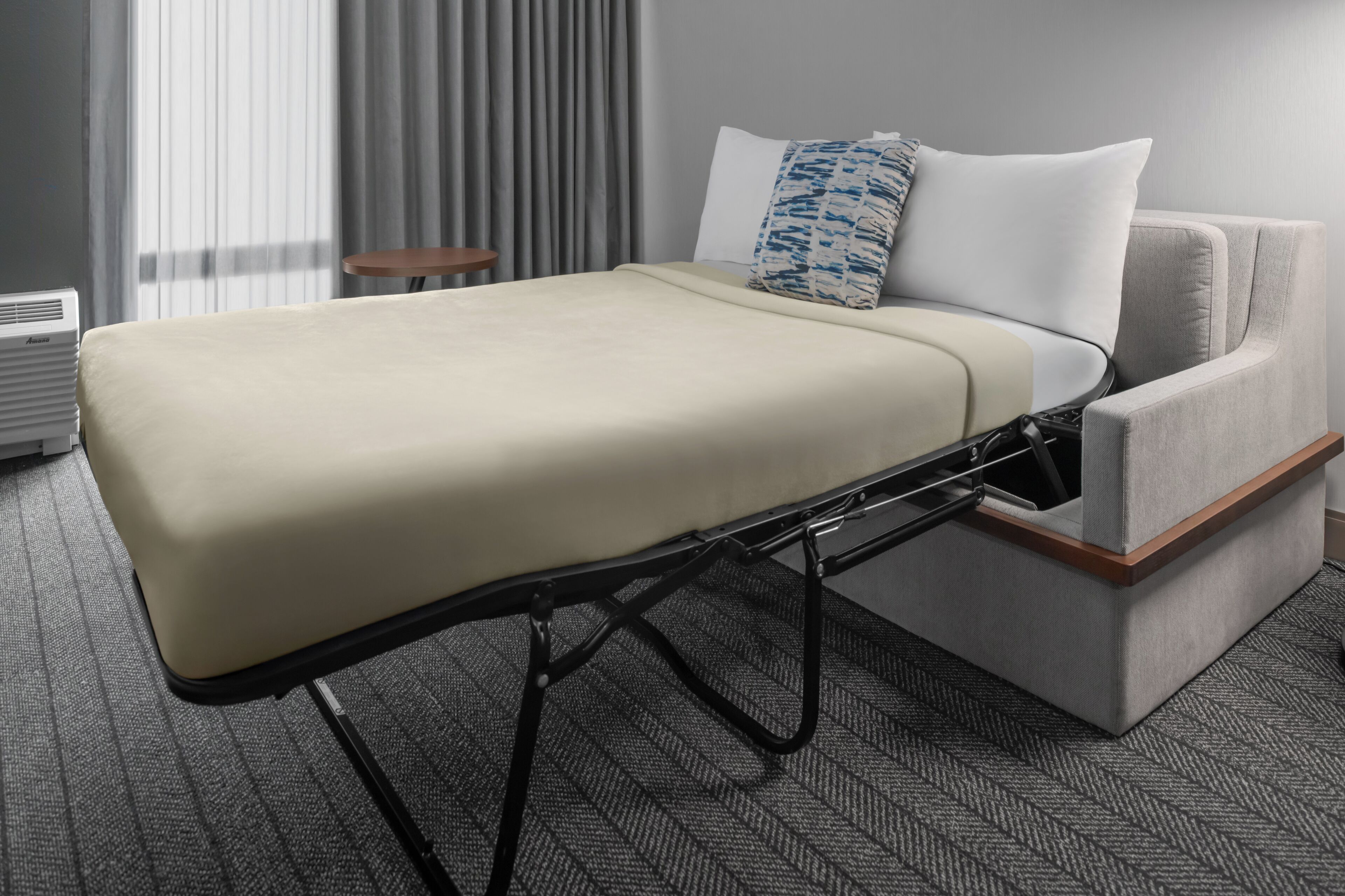 Una cama compacta con colchón y cabecera, una mesita de madera pequeña y una silla tapizada de color gris.