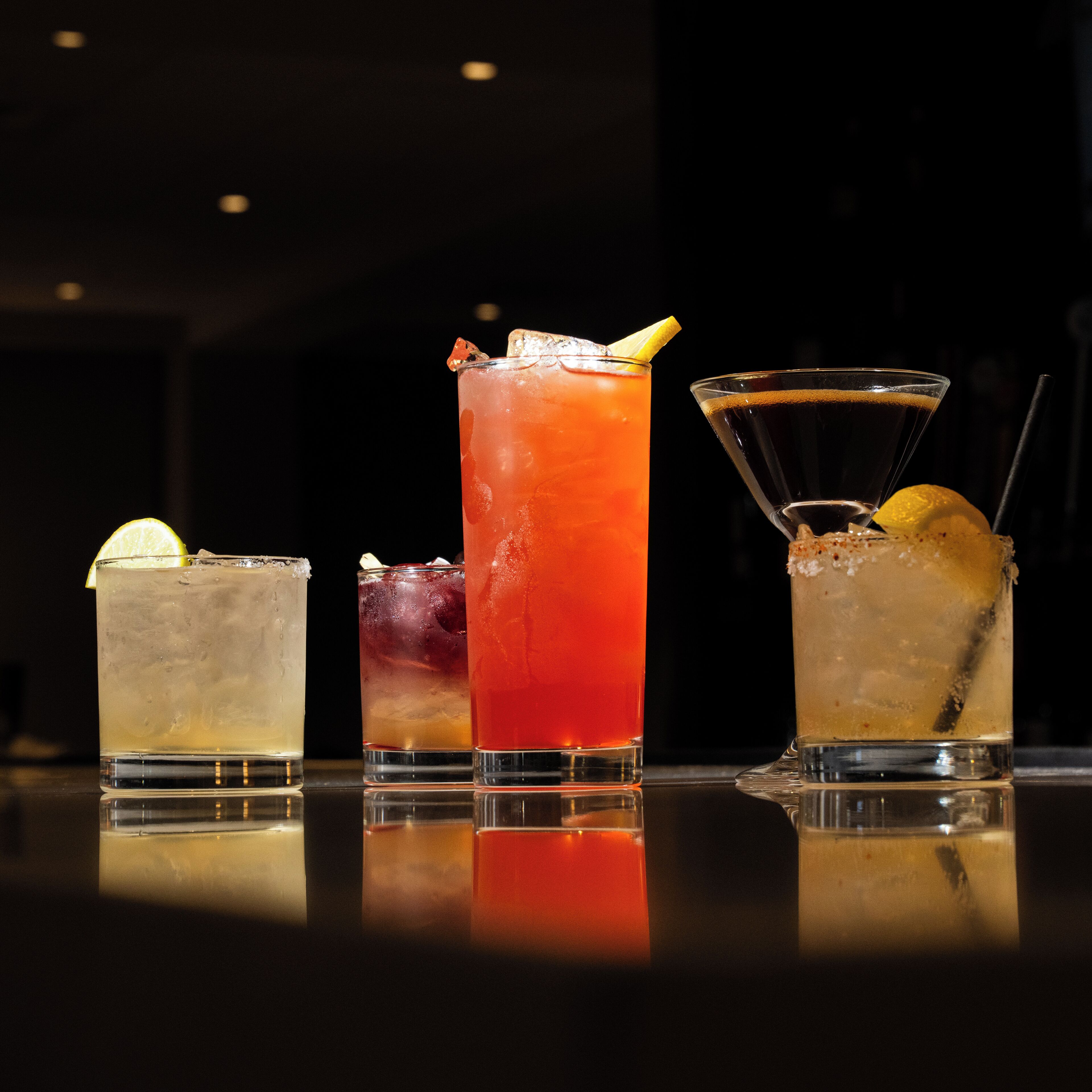 Four glasses of different beverages on a reflective surface.