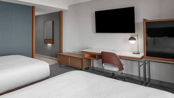 A hotel room with two beds, a desk with a chair, a flat-screen TV, a bedside lamp, and a large mirror.