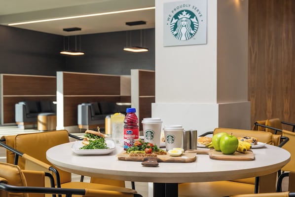 Un comedor con una mesa dispuesta para una comida, que incluye un sándwich, ensalada y fruta. Hay tazas Starbucks y una botella de jugo de arándano en la mesa. La habitación tiene muebles modernos y un logotipo de Starbucks en la pared.