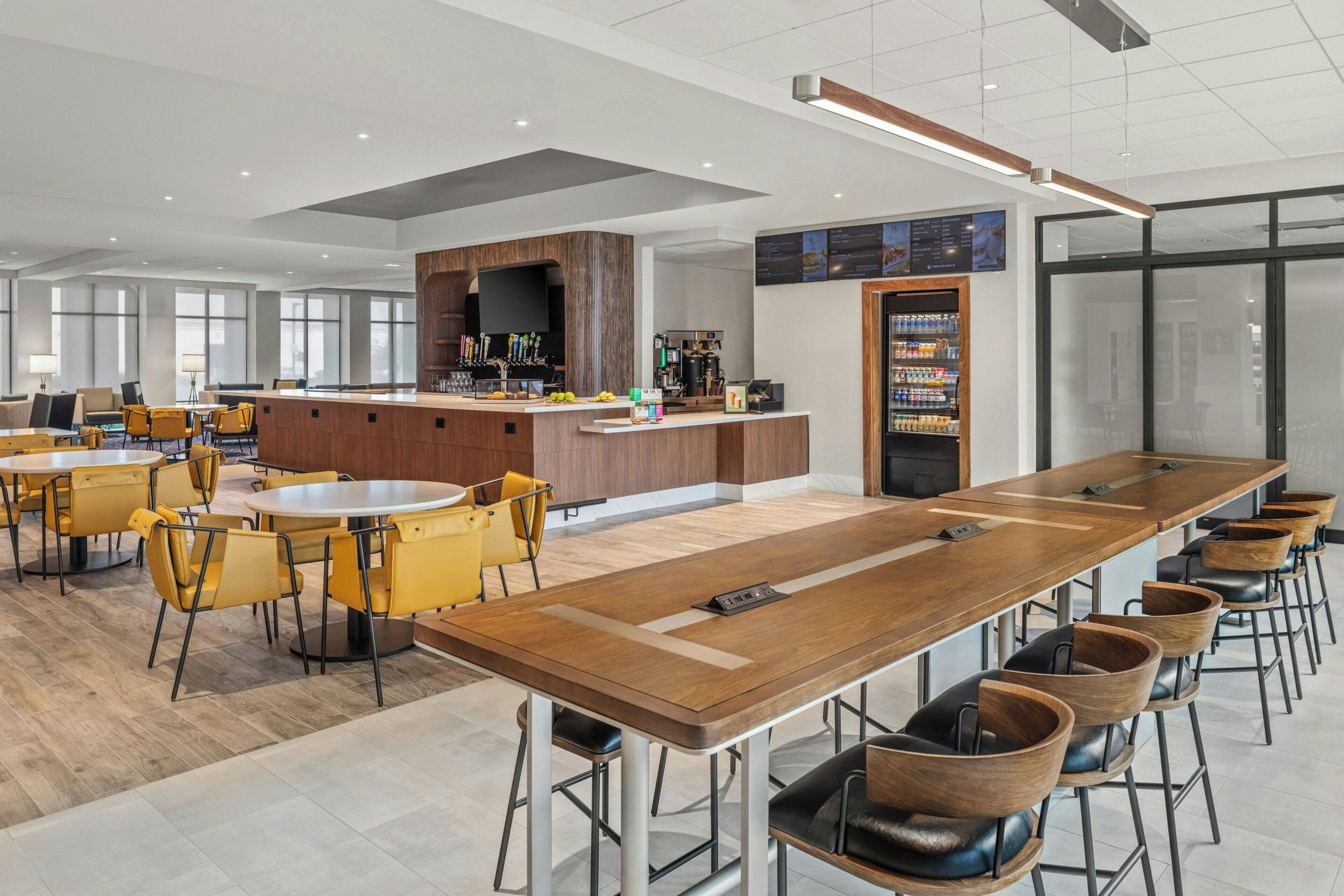 Un moderno vestíbulo de hotel con un largo mostrador de recepción de madera, varias zonas de estar con sillas amarillas y negras, y una zona de bar con una pantalla de televisión.