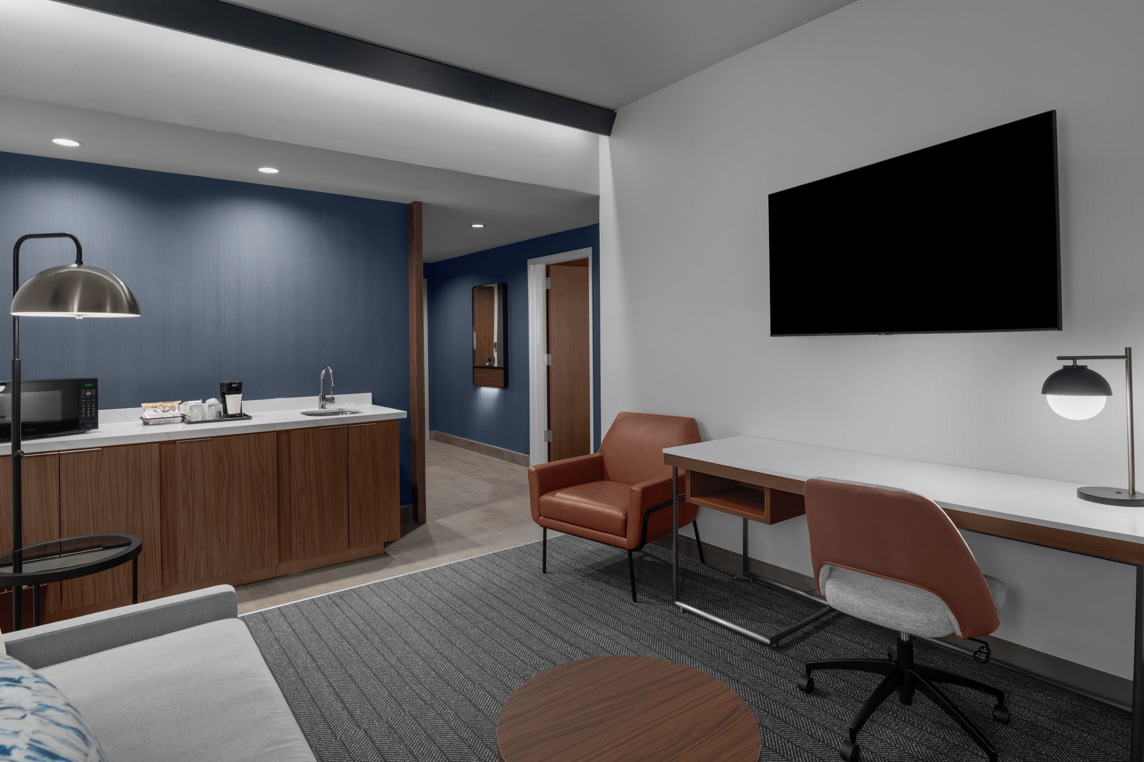 A modern hotel room with a kitchenette, a desk, a chair, a bed, and a TV mounted on the wall.