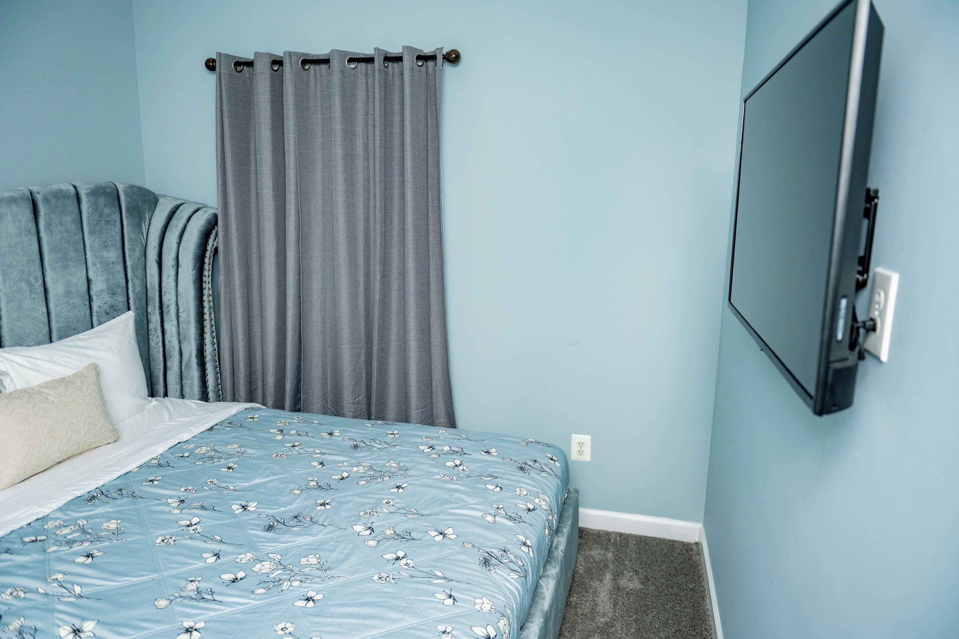 Quarto com cama, cortinas cinzas, televisão montada e parede azul claro.