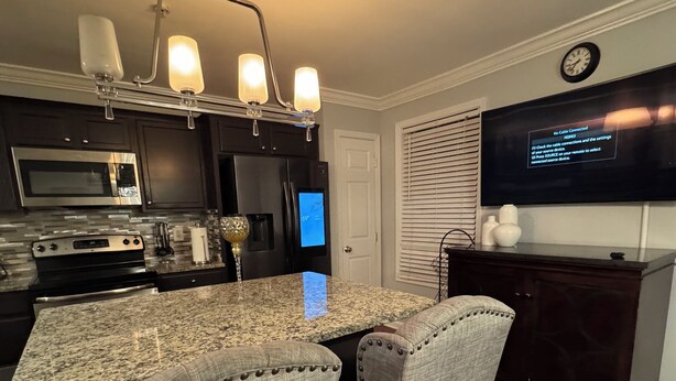 A modern kitchen with a granite countertop, stainless steel appliances, and a built-in oven. There is a dining area with a patterned rug and upholstered chairs.