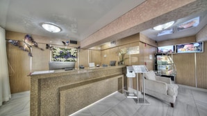 Zona de recepción de un hotel con mostrador de granito, decoración floral en la pared, un sofá y una pantalla de televisión mostrando imágenes.