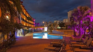 Zona de piscina de hotel con sillones de descanso, palmeras y un letrero que indica el Hotel Royal Beach.