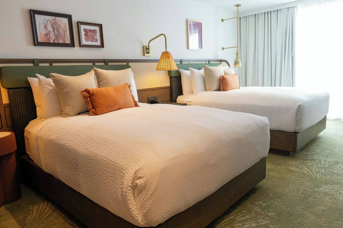 Une chambre d’hôtel avec deux lits, une tête de lit verte, une grande fenêtre avec des rideaux et des œuvres d’art encadrées accrochées au mur.