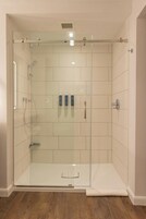 Una ducha con paredes de vidrio, pared de azulejos blancos, suelo de madera y una toalla en el piso.