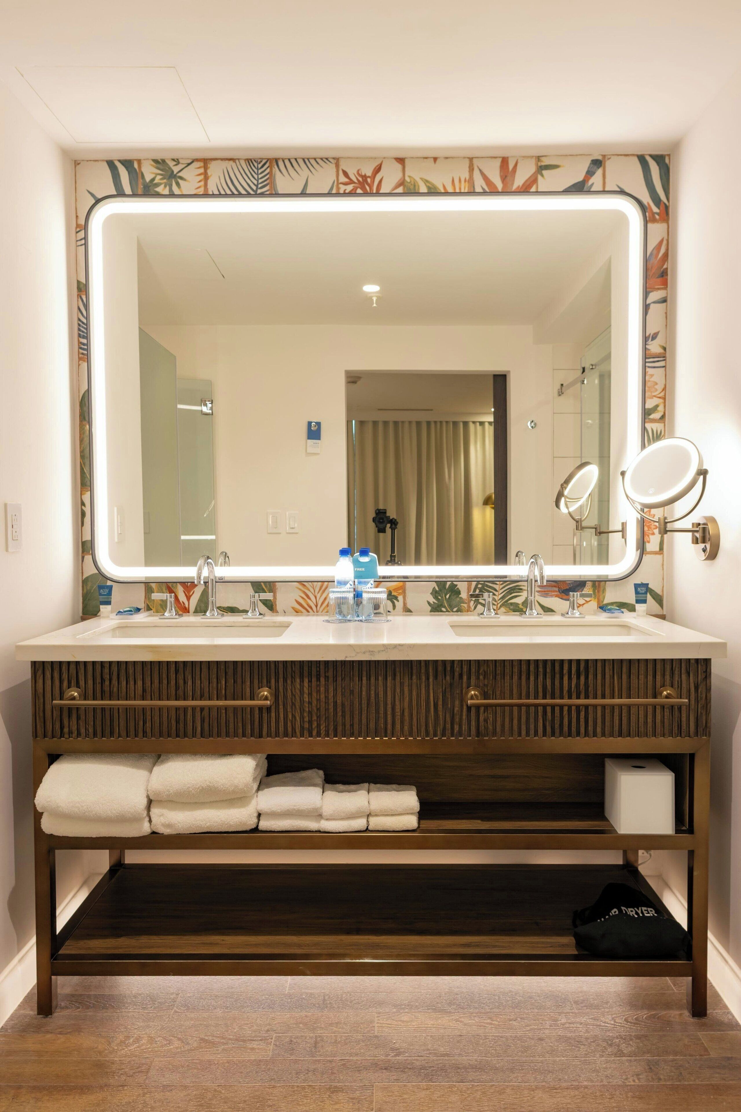 A bathroom with a large mirror, two sinks, and a vanity with shelves.