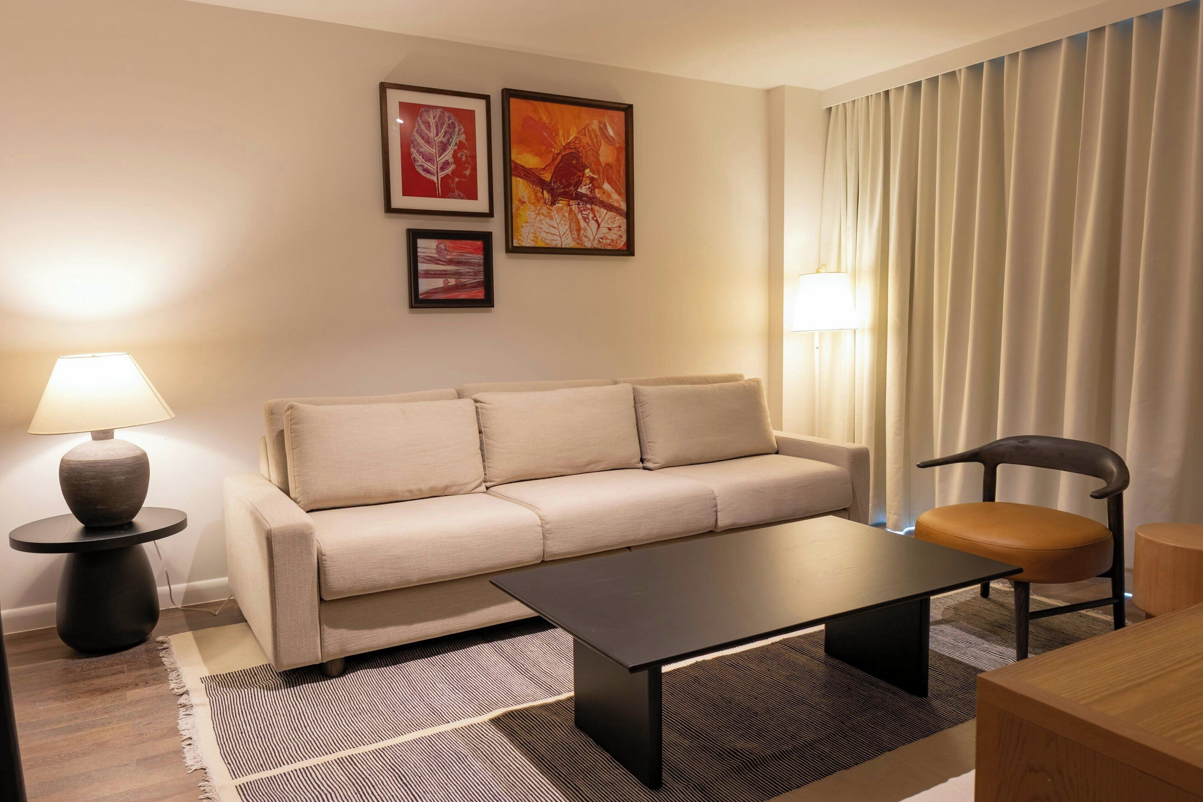 A modern living room with a sofa, a coffee table, a chair, and a lamp. There are three framed artworks on the wall.