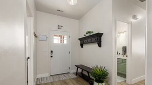 Un couloir avec une porte blanche portant l’inscription « SORTIE », un banc en bois sombre, une plante en pot et une étagère fixée au mur au-dessus du banc.