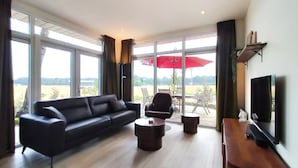 A modern living room with a black sofa, a coffee table, and a TV on a wooden stand. Large windows offer a view of the outdoors. - 2 bedroom cozy home in Hellendoorn (Hellendoorn)