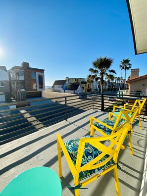 Een terras op het dak met gele stoelen en een blauwe tafel, met uitzicht op een strand met palmbomen en moderne gebouwen.