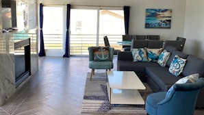 Un vaste espace de vie doté d’un parquet en point de Hongrie, d’un canapé d’angle agrémenté de coussins à motifs, d’un fauteuil bleu et d’un téléviseur à écran plat fixé sur un mur en marbre.